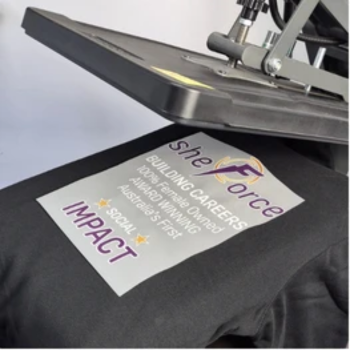 Heat press applying a "SheForce" transfer reading "Building Careers, Social Impact" onto a black garment.